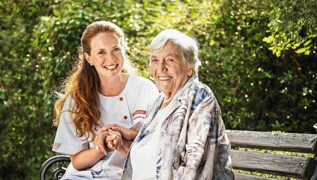 Pflegekraft sitzt mit Seniorin auf einer Bank