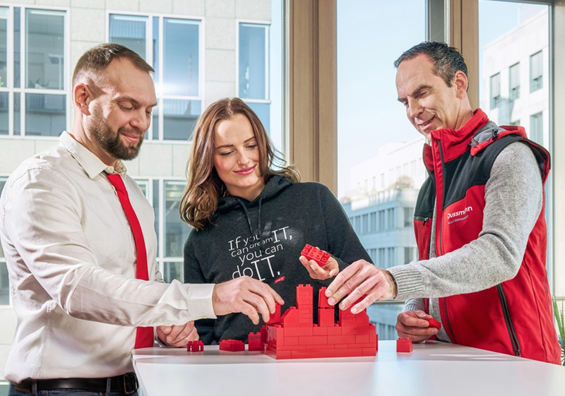 Drei Personen bauen gemeinsam einen Turm aus roten Bausteinen